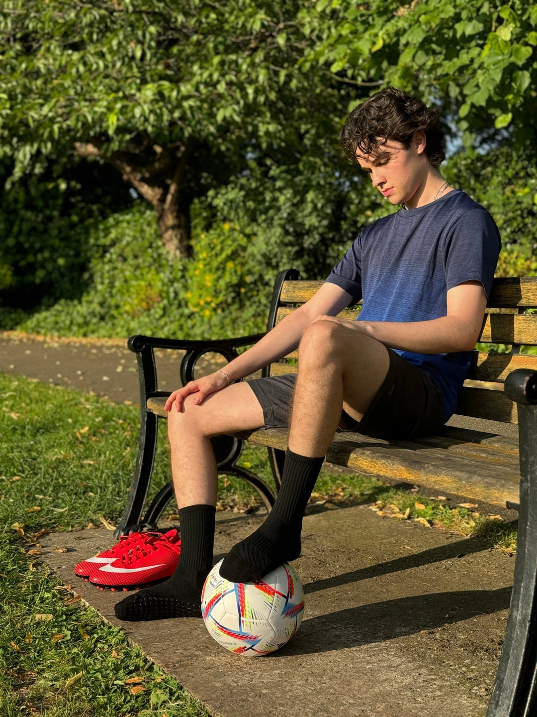 Athlete wearing Fitknitsocks black grip socks, sitting on a park bench with a football and red boots