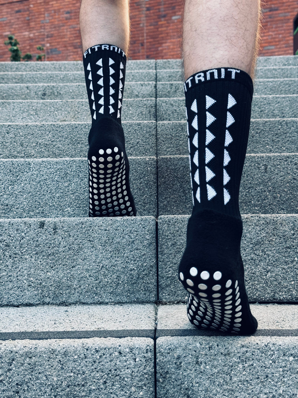 A person wearing FitKnitSocks black grip socks with a white geometric pattern and anti-slip soles, walking up a set of concrete stairs